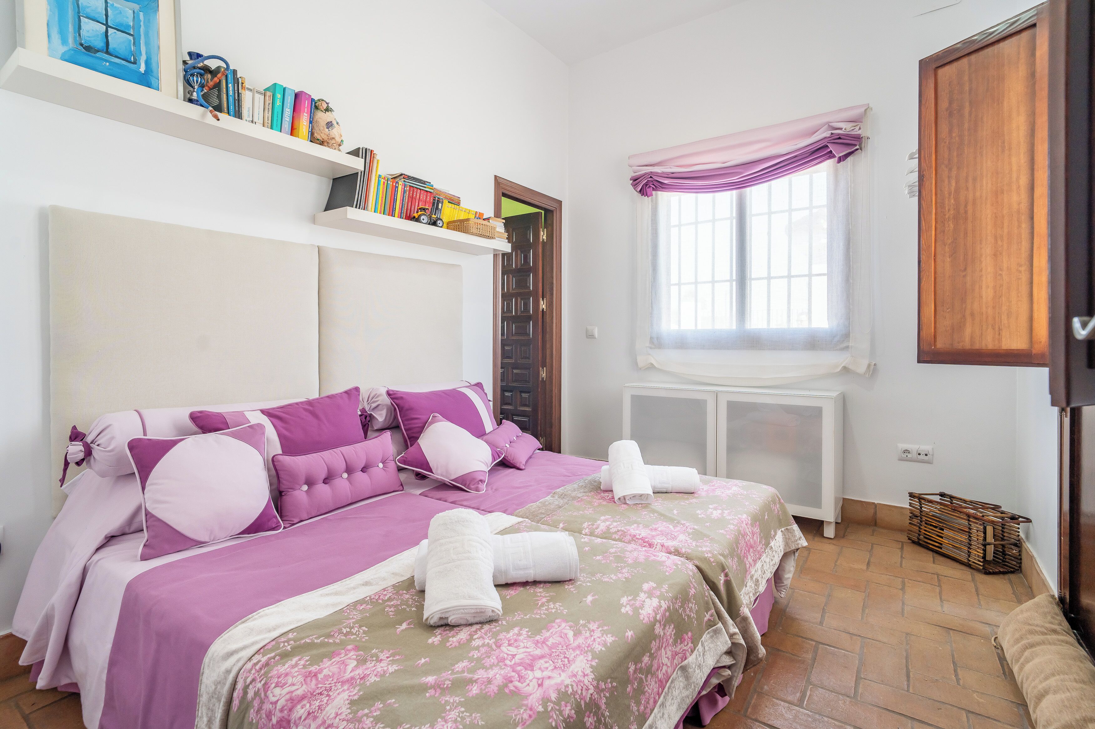 Una camera da letto con due letti, una finestra con tende, un termosifone e uno scaffale con libri e giocattoli.