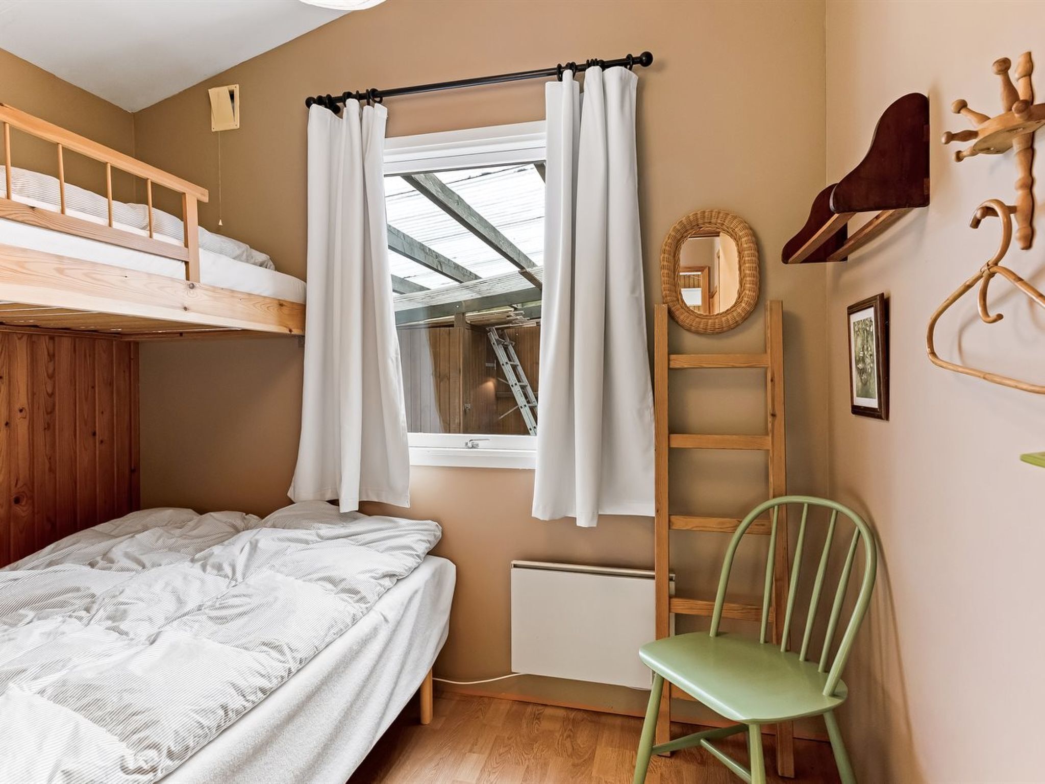 A bedroom with a bunk bed, a window with white curtains, a wooden ladder, a green chair, and a wall-mounted mirror.