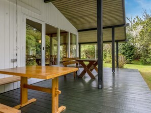 An outdoor wooden deck with a picnic table and benches, surrounded by glass doors leading to a lush garden.