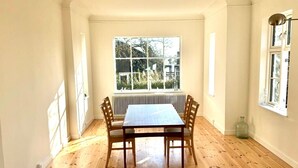 Een kamer met een houten vloer, een tafel met vier stoelen, een radiator en een raam met uitzicht op bomen en gebouwen.