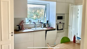 Een compacte keuken met witte kastjes, een raam met uitzicht op bomen, een magnetron en een waterkoker.