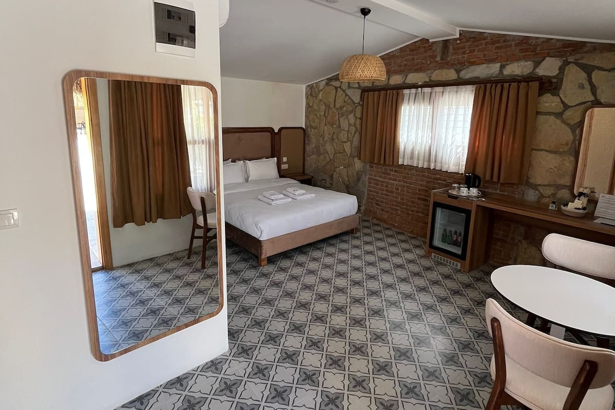A hotel room with a bed, a small table, a chair, a mirror, a television, and a stone wall.