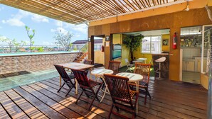 An outdoor patio with wooden tables and chairs, a view of the pool, and a restaurant area with a counter and stools. - Hotel O My Home@Bali (Kuta)