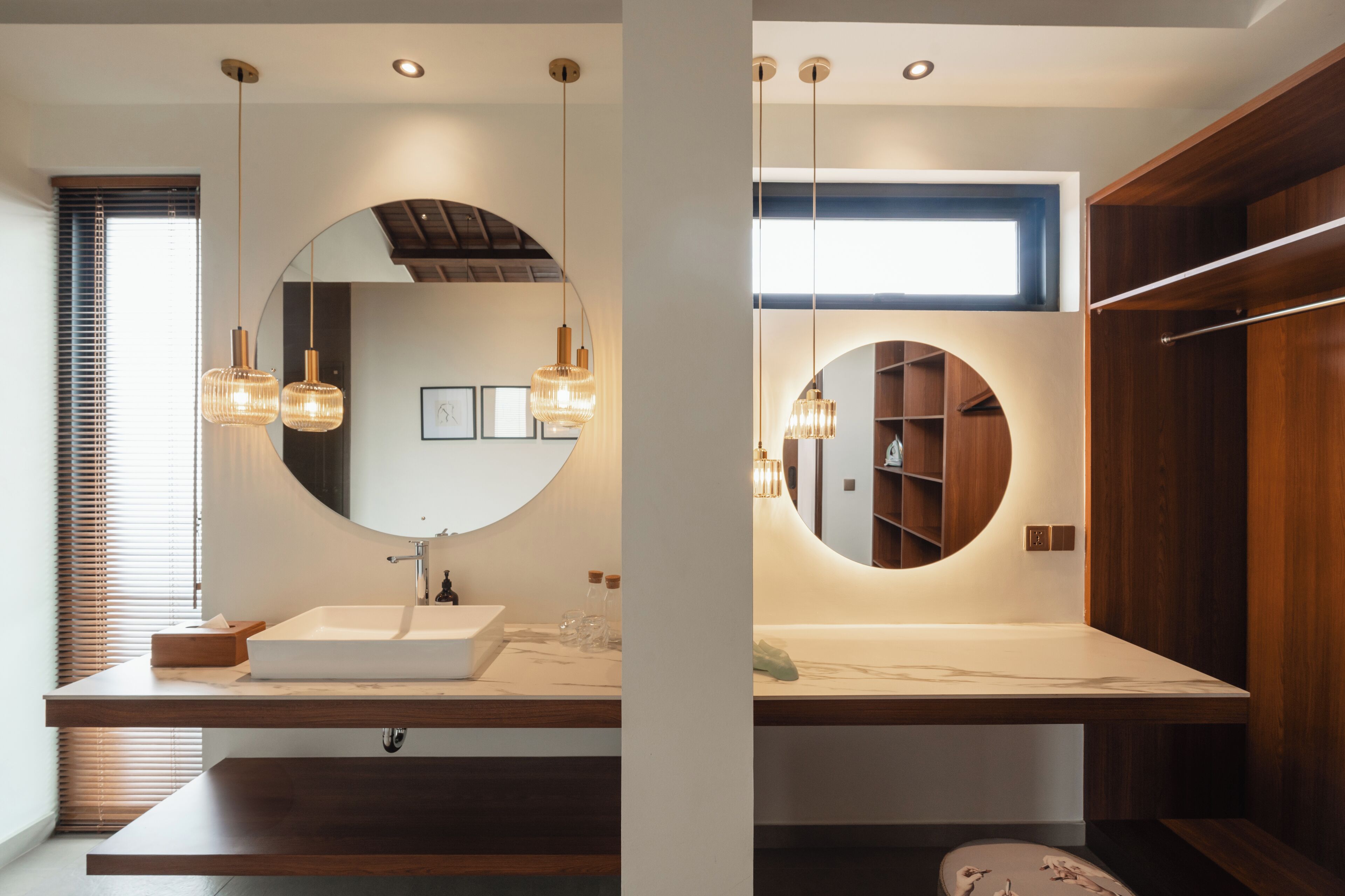 A modern bathroom with a large round mirror, wooden vanity, and pendant lighting.