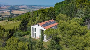 A modern house with a red roof, surrounded by dense green trees and hills. - Elegant Villa with Garden - Perfect for Groups (Potenza Picena)