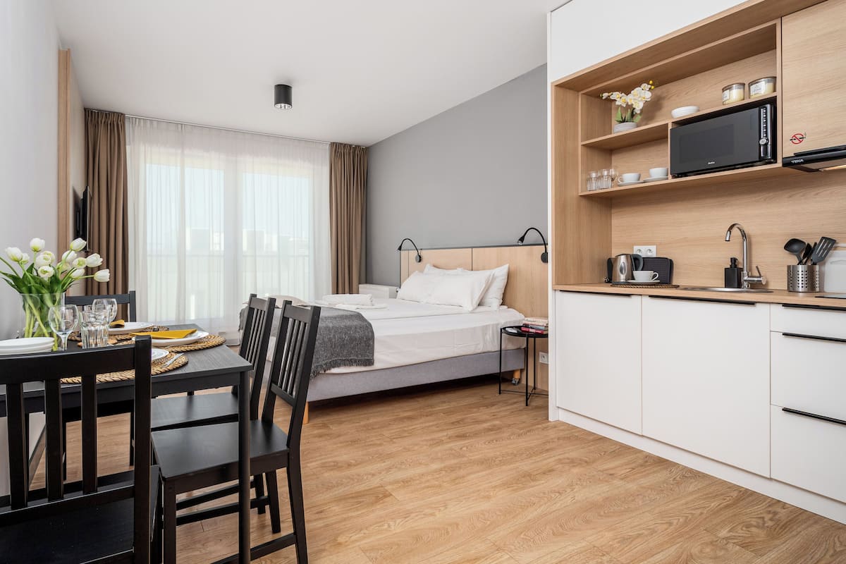 A modern hotel room with a bed, a dining table with chairs, a kitchen area with a microwave, and a window with curtains.