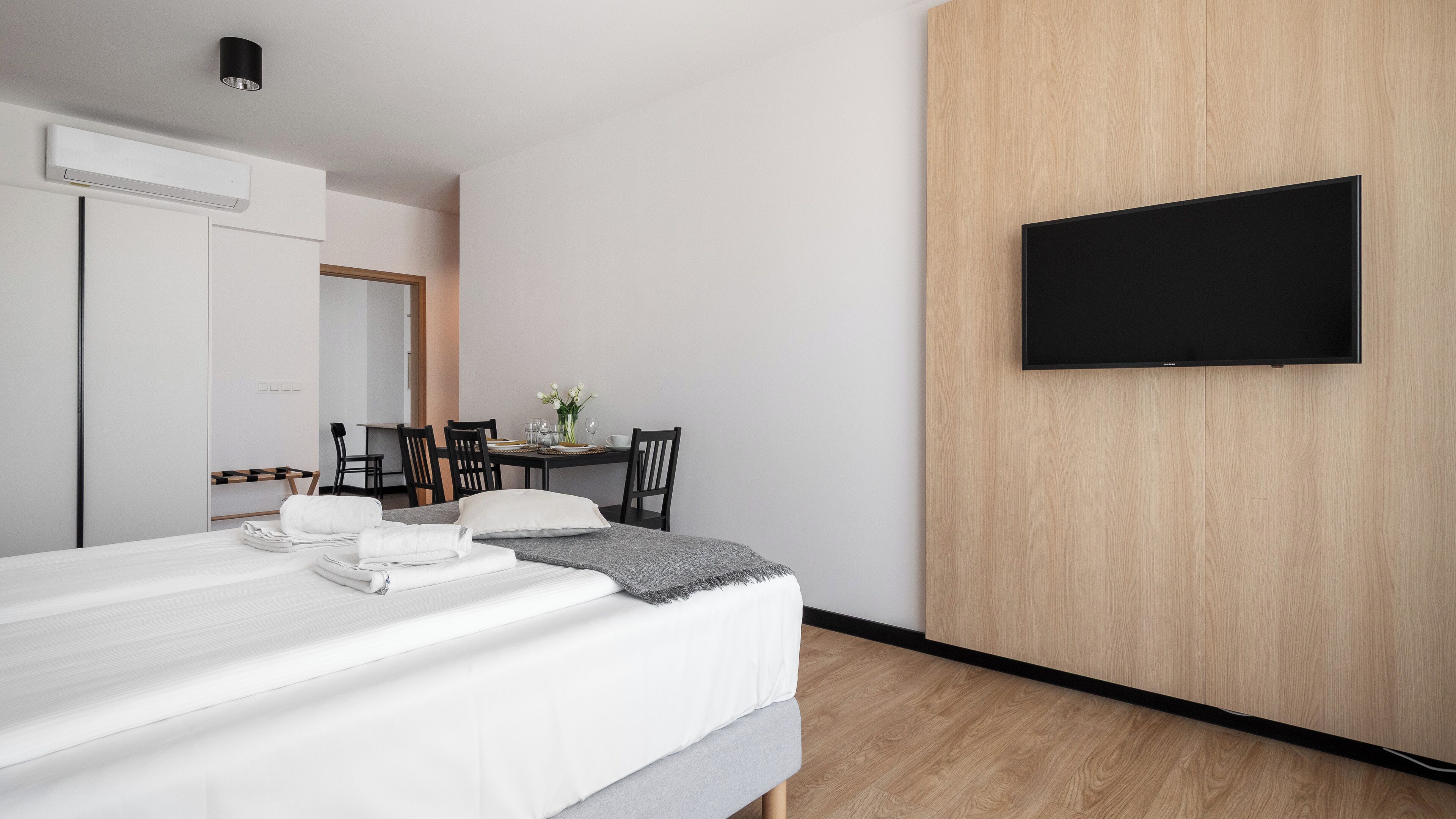 A modern hotel room with a bed, a TV mounted on a wooden wall, and a dining area with chairs and a table.