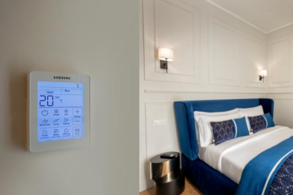 A room with a Samsung thermostat displaying 20°C, a bed with blue bedding, and a wall-mounted lamp.