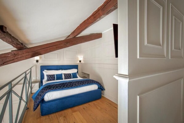 A bedroom with a blue bed, wooden beams, white walls, and a wooden floor.
