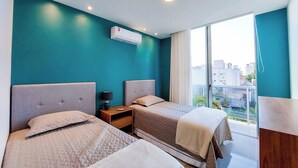 Habitación de hotel con dos camas, una mesita de noche, un equipo de aire acondicionado y vistas a la ciudad a través de amplias ventanas.