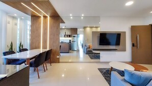 Una sala moderna con televisor de pantalla plana montado en la pared, un comedor con mesa larga y sillas, y una cocina al fondo.