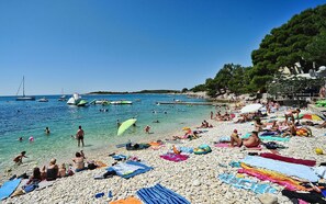 A crowded beach with people sunbathing and swimming, surrounded by boats and a rocky shoreline.