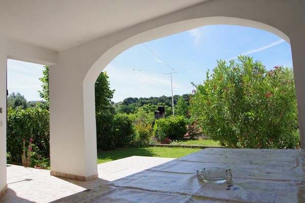 A covered outdoor dining area with a view of a lush garden and a clear sky.