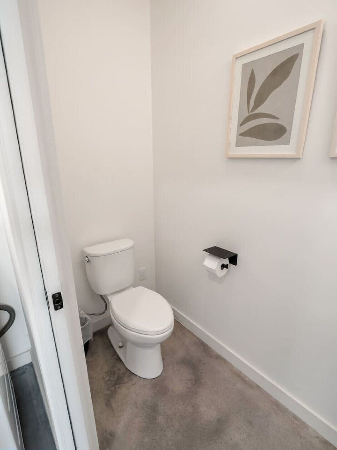 Un baño pequeño y blanco con un inodoro, un rollo de papel higiénico en su soporte y una obra de arte abstracto enmarcada en la pared.