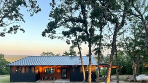 Een moderne cabin met een metalen dak, omgeven door bomen en een grindpad.