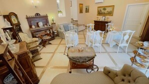 A spacious living room with a large sofa, a coffee table, and a dining area with white chairs. There is a wooden cabinet and a clock on the wall.
