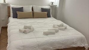 Un lit bien fait, avec du linge de lit blanc et des serviettes pliées, une tête de lit en bois et deux lampes de chevet posées sur des tables de nuit blanches.
