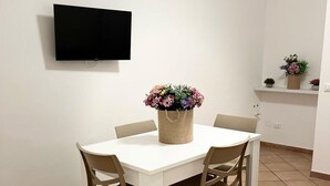 Un coin repas avec une table blanche et quatre chaises, un téléviseur à écran plat fixé au mur et une plante en pot posée sur la table.