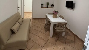 Un espace de vie compact comprenant un canapé beige, une table à manger avec des chaises, un petit téléviseur fixé au mur et une fenêtre donnant sur un paysage agréable.