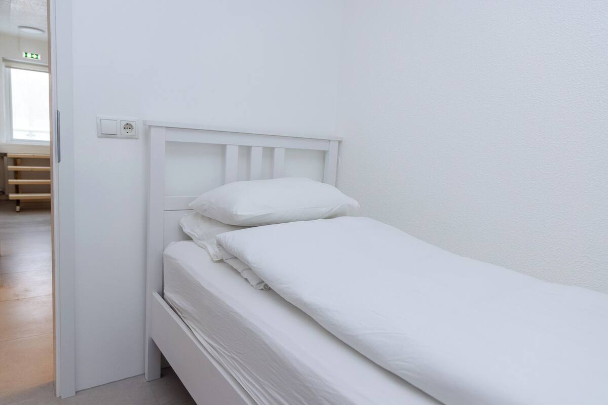 Een wit eenpersoonsbed met hoofdeinde, nachtkastje en een stopcontact aan de muur.