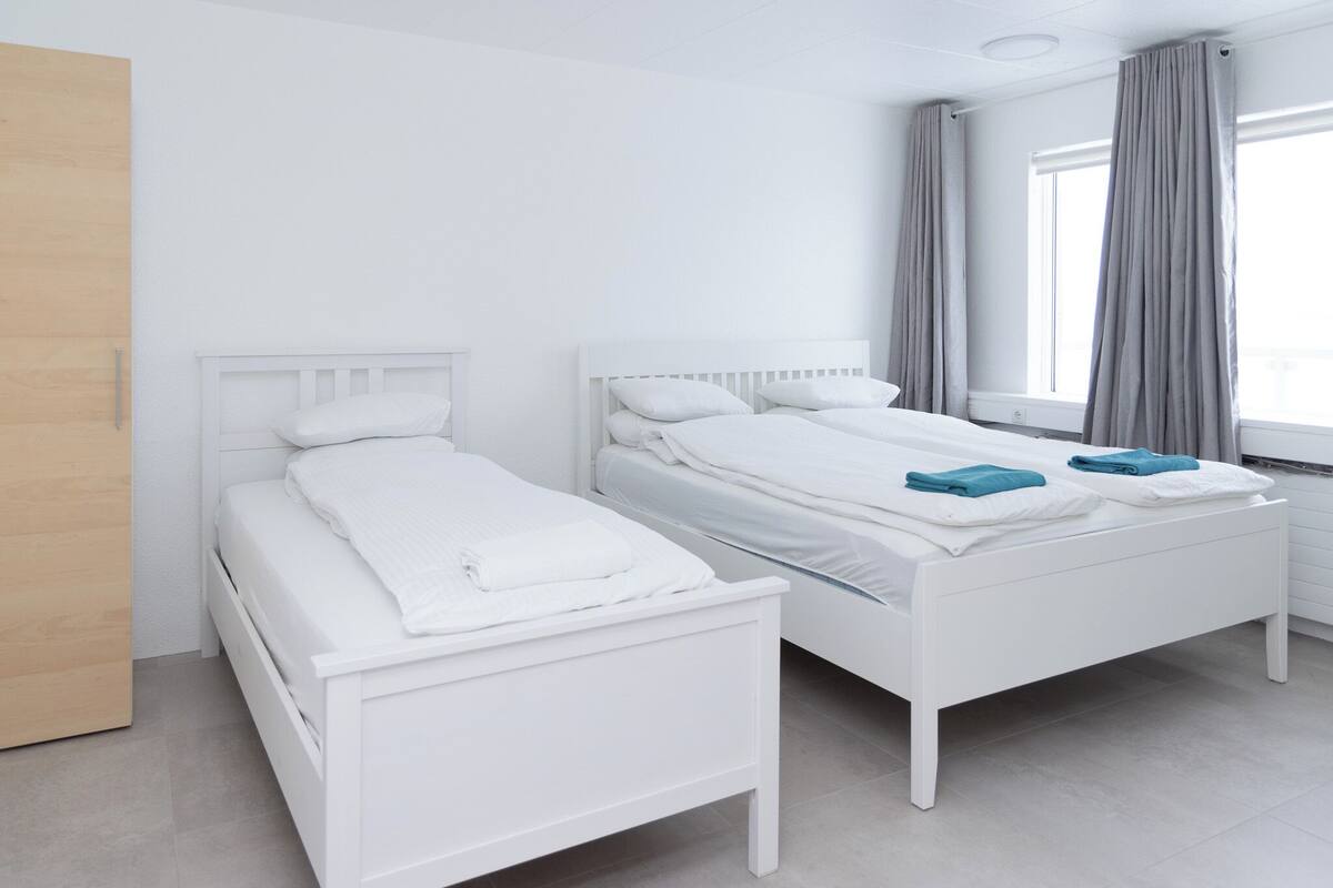 Een moderne hotelkamer met twee witte bedden, een houten deur en een raam met grijze gordijnen.