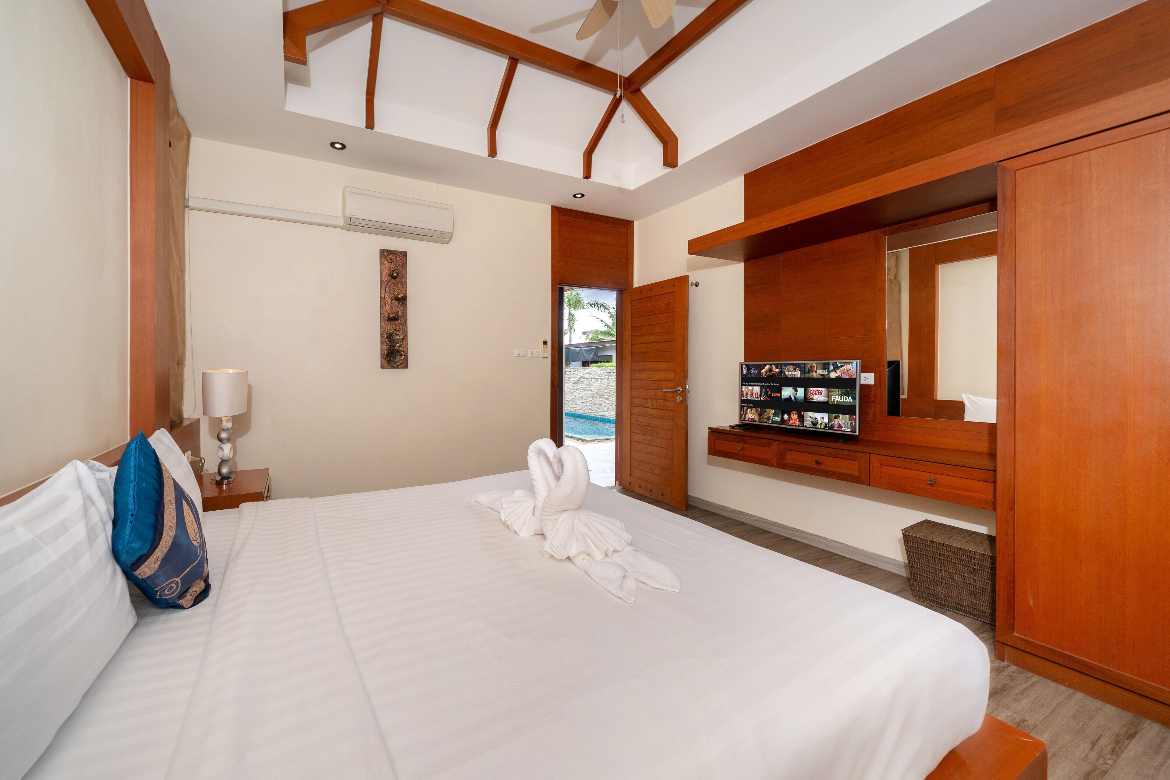 A hotel room with a large bed, wooden headboard, a wardrobe, a vanity with a mirror, and a wall-mounted TV.