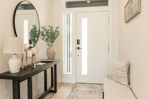 Un couloir avec une console sombre, un miroir, une plante en pot, une porte surmontée d’un luminaire et un petit tapis.