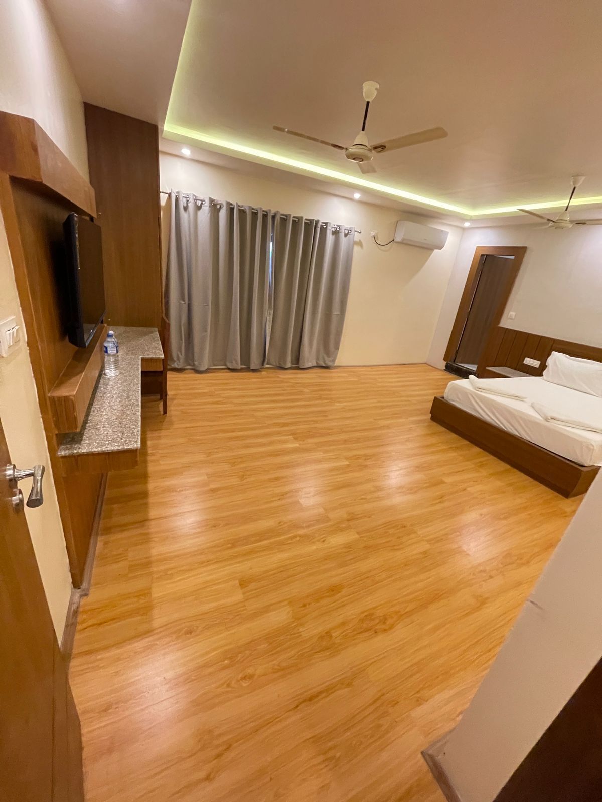 Quarto de hotel com piso de madeira, uma cama, televisão em um suporte, uma mesinha com uma garrafa de água e um ventilador de teto.