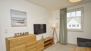 A modern living room with a wooden cabinet, a flat-screen TV, a lamp, and a view of a house through a window. - Beachfront vacation rental with a balcony, access to the pool and sauna - Rex Rugia (Mönchgut OT Lobbe)