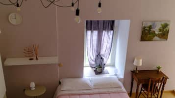 Quarto com cama, mesa de cabeceira, abajur e janela com cortinas.