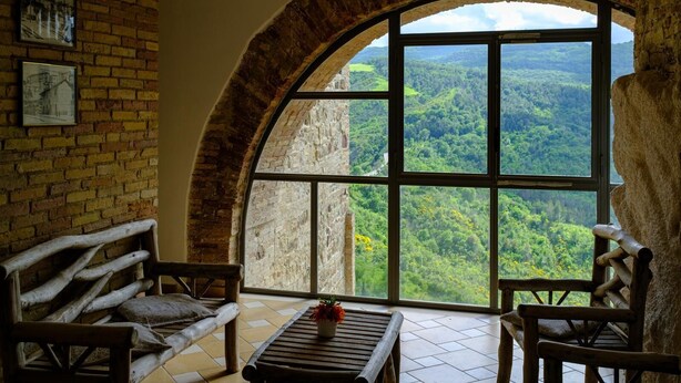 A rustic room with a large arched window offering a view of a forested valley. There is a wooden bench and a small table with a flower vase. - Il castello di atessa  (Atessa)