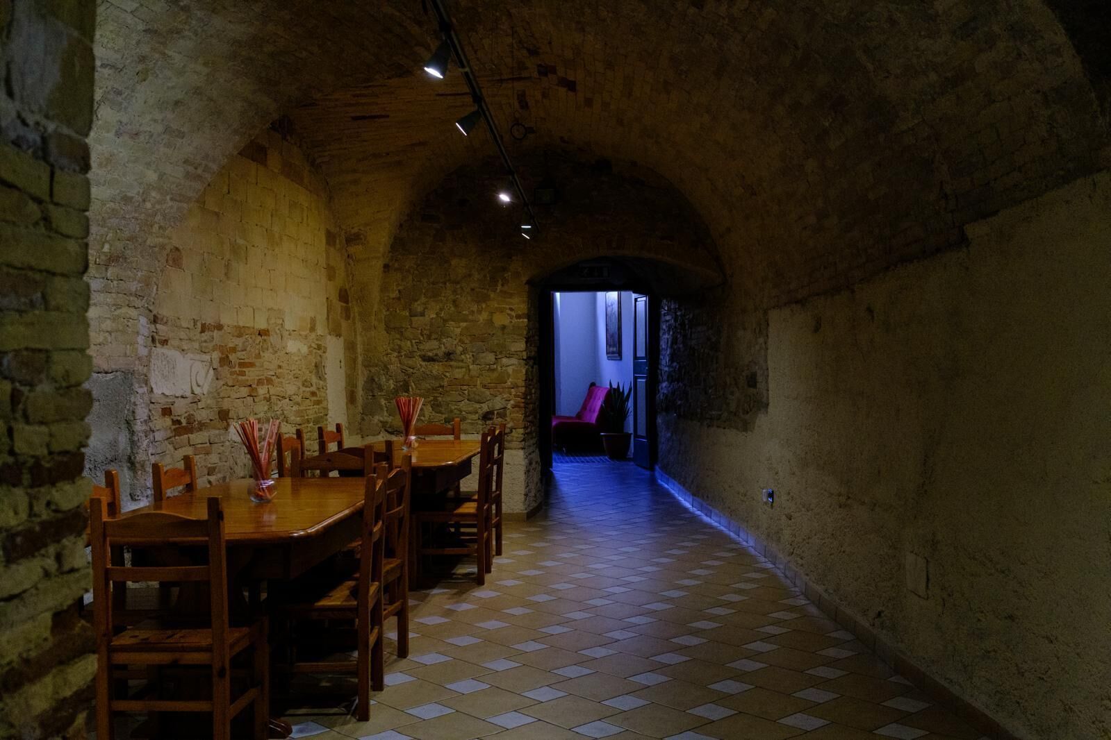Un estrecho pasillo con paredes de piedra y suelo de baldosas que conduce a una habitación con una silla roja y una mesa.