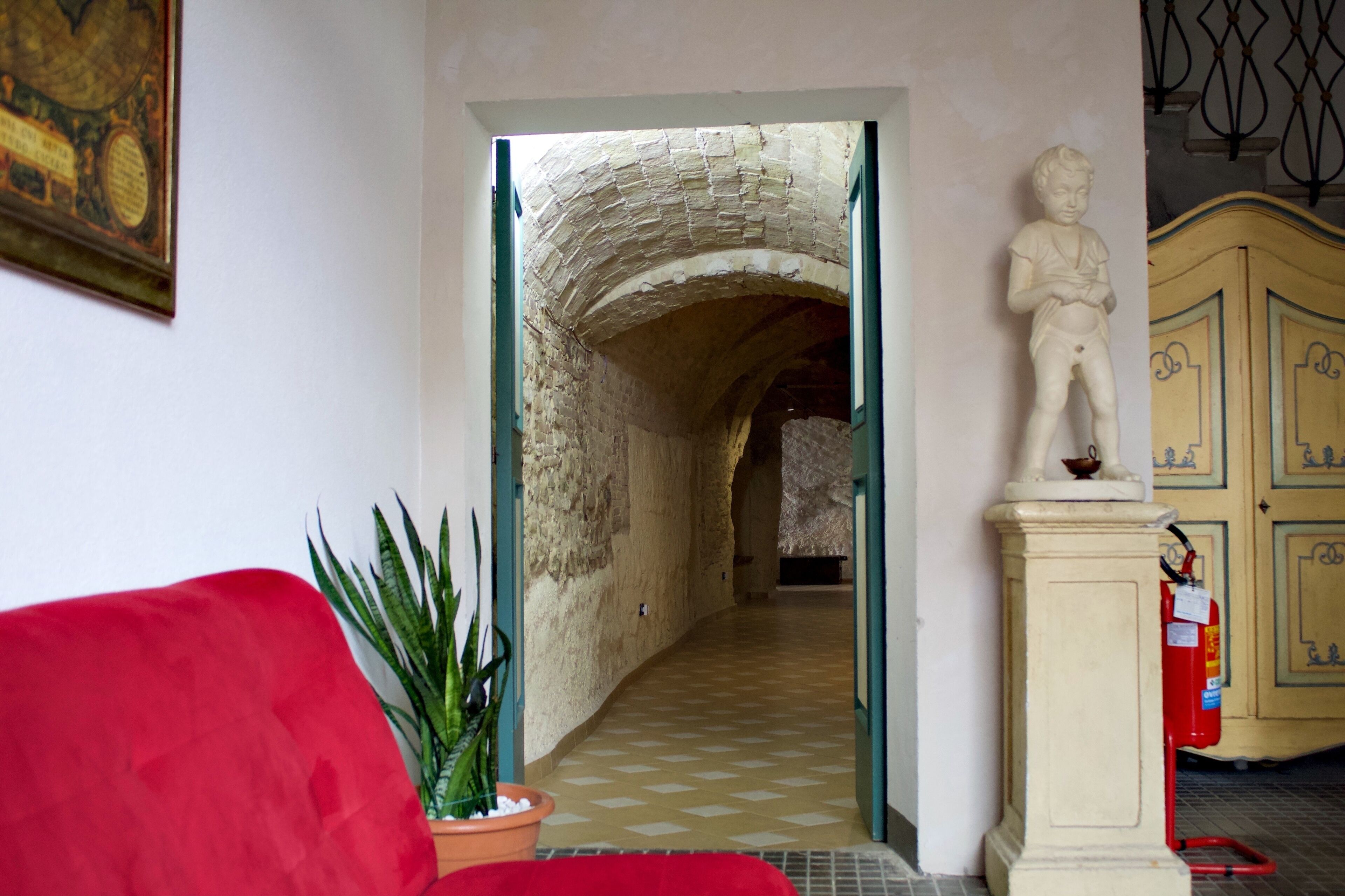 A hallway with a stone archway leading to another room, a red sofa, a potted plant, and a statue on a pedestal.
