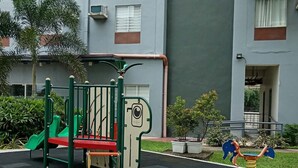 Um parquinho com escorrega, estrutura de escalada e balanços, cercado por um edifício com sacadas e janelas.