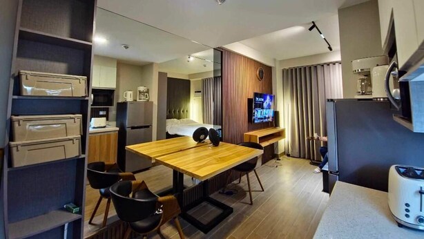 Habitación de hotel moderna con una mesa de comedor de madera, sillas negras, televisor, zona de cocina y estanterías empotradas.