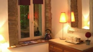 Una stanza con parete in pietra, una cassettiera in legno, uno specchio, una finestra con persiane, un tavolo con una lampada e una sedia.