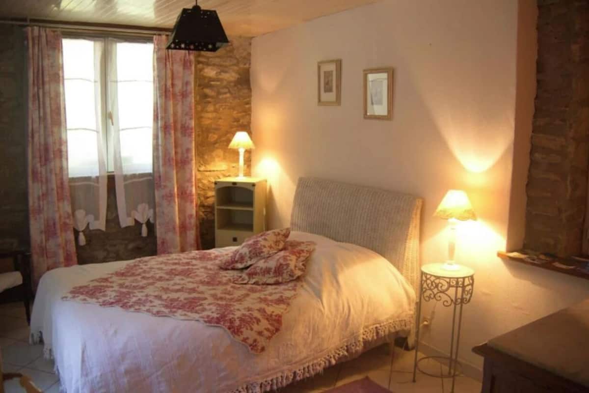 A cozy bedroom with a bed, two bedside lamps, a stone wall, and a window with curtains.