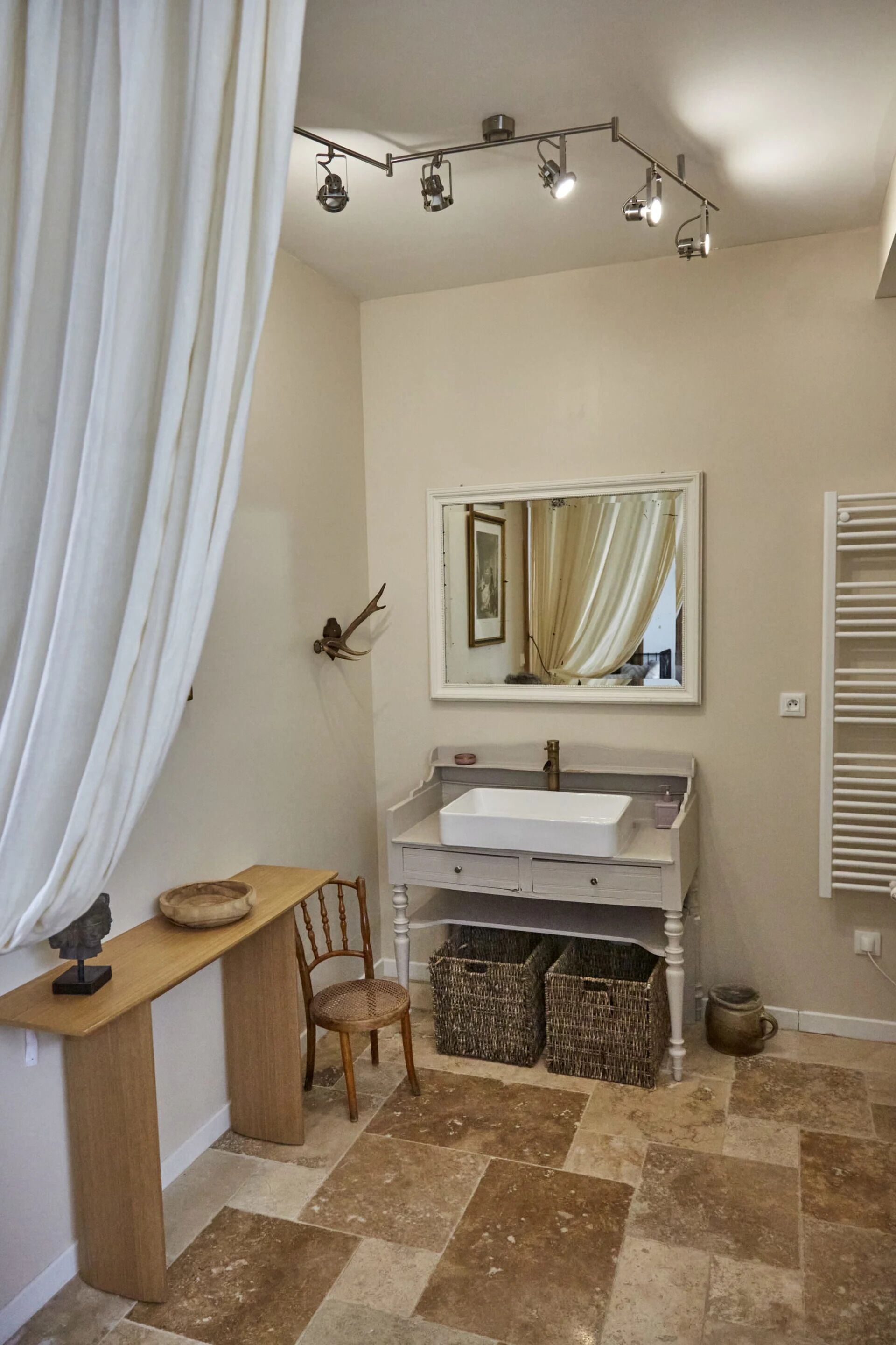 A bathroom with a white sink, mirror, and ceiling lights. There is a wooden chair and two wicker baskets on the floor.