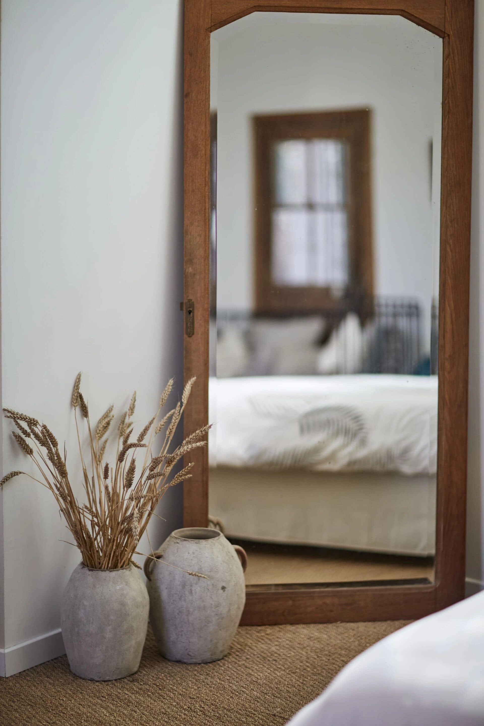 Une chambre avec une porte en bois, deux vases gris contenant des plantes séchées et un lit avec une literie blanche.