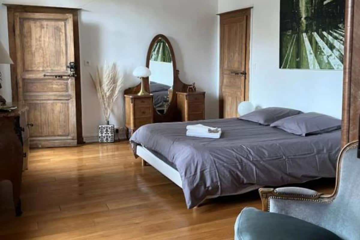 A bedroom with a wooden floor, a bed with grey bedding, a wooden wardrobe, a dresser, a mirror, and a painting on the wall.
