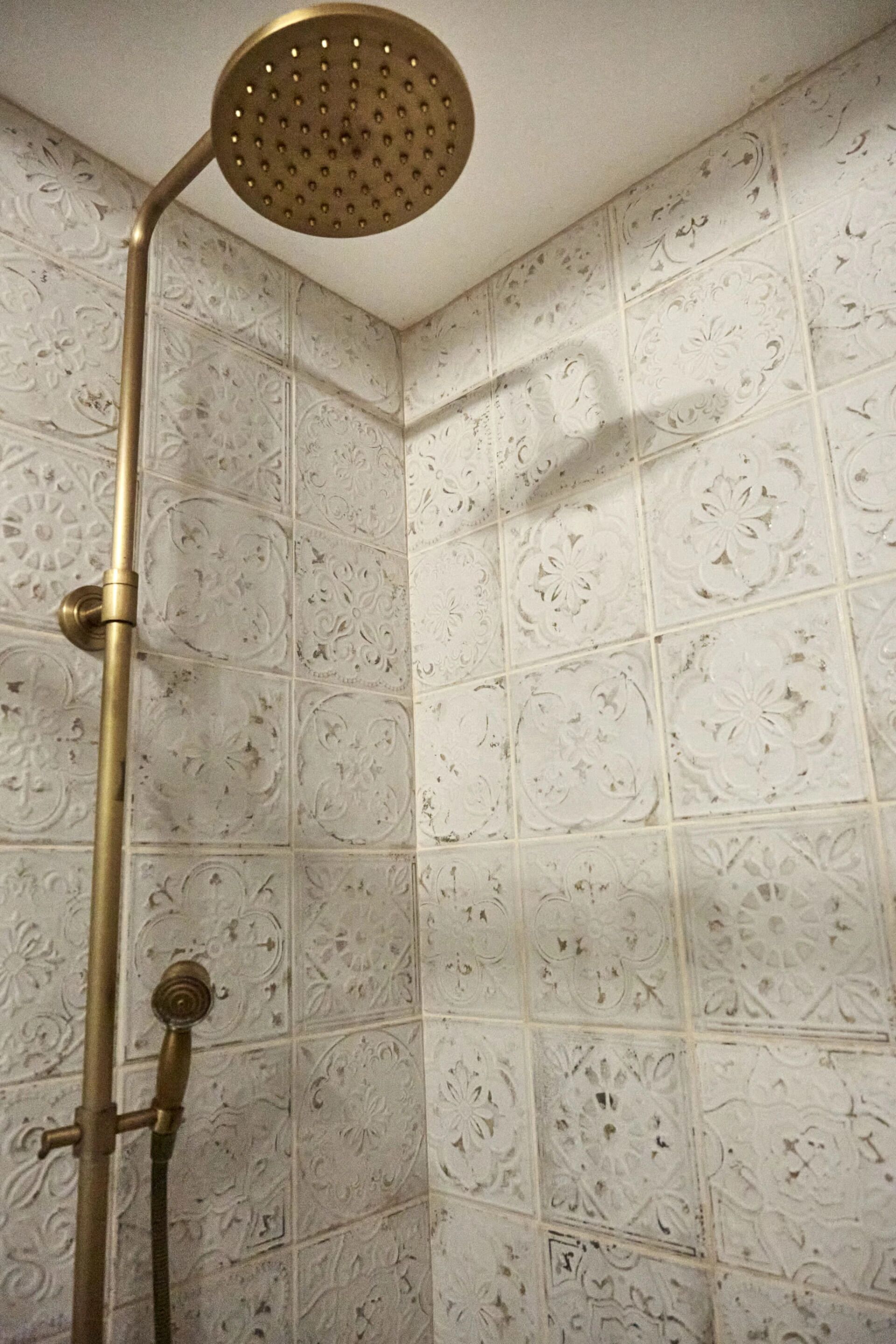 A shower with a gold showerhead and brass fixtures against a wall with ornate, embossed tiles.