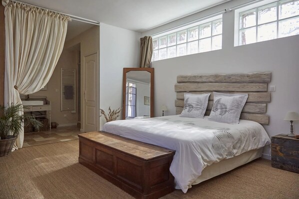 A bedroom with a wooden headboard, a large bed with white bedding, a wooden nightstand, a mirror, and a window with curtains. - L'ÉVIDENCE (Rugles)