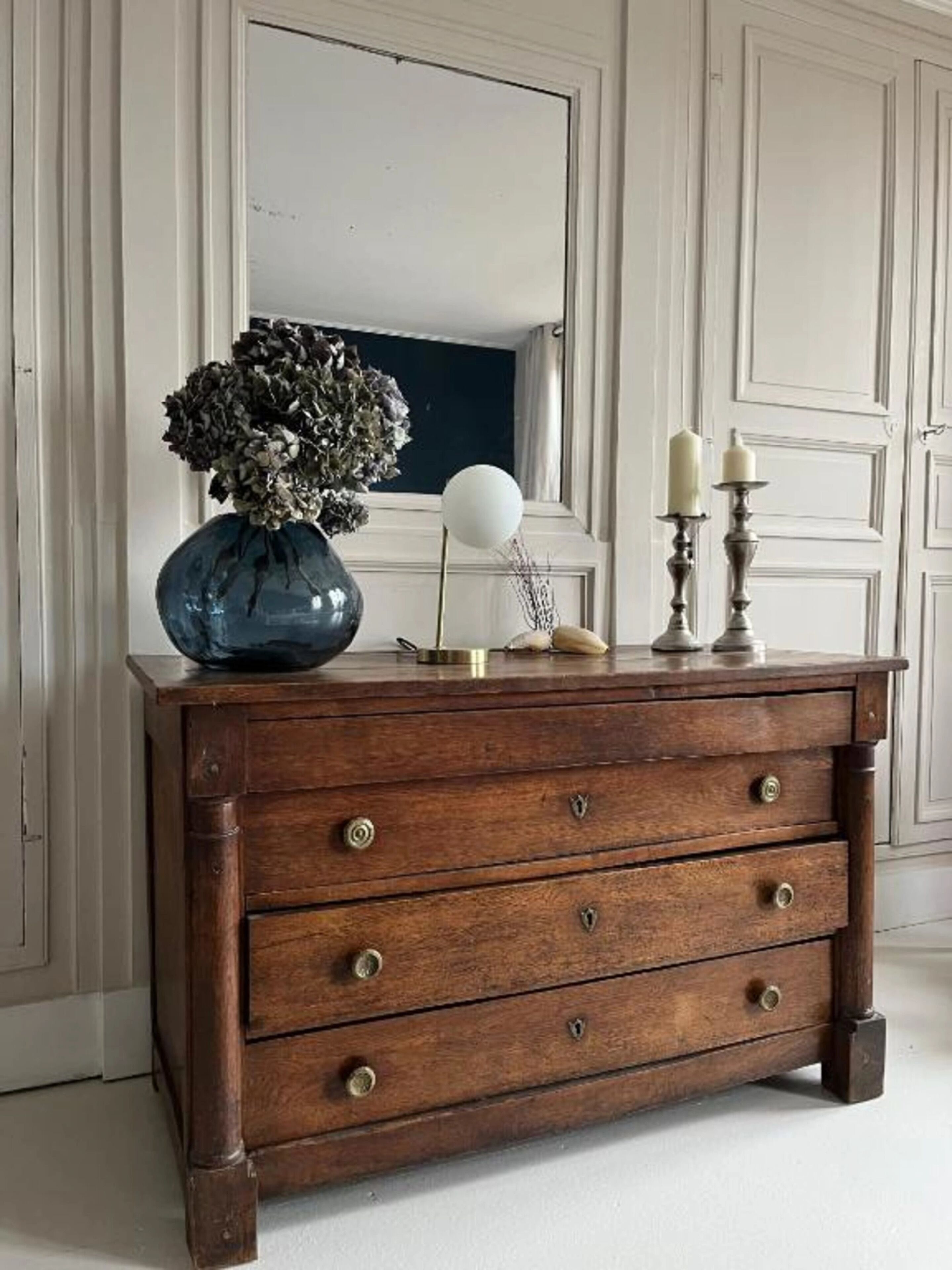 A wooden dresser with three drawers, a vase with dried flowers, and two candles on top.
