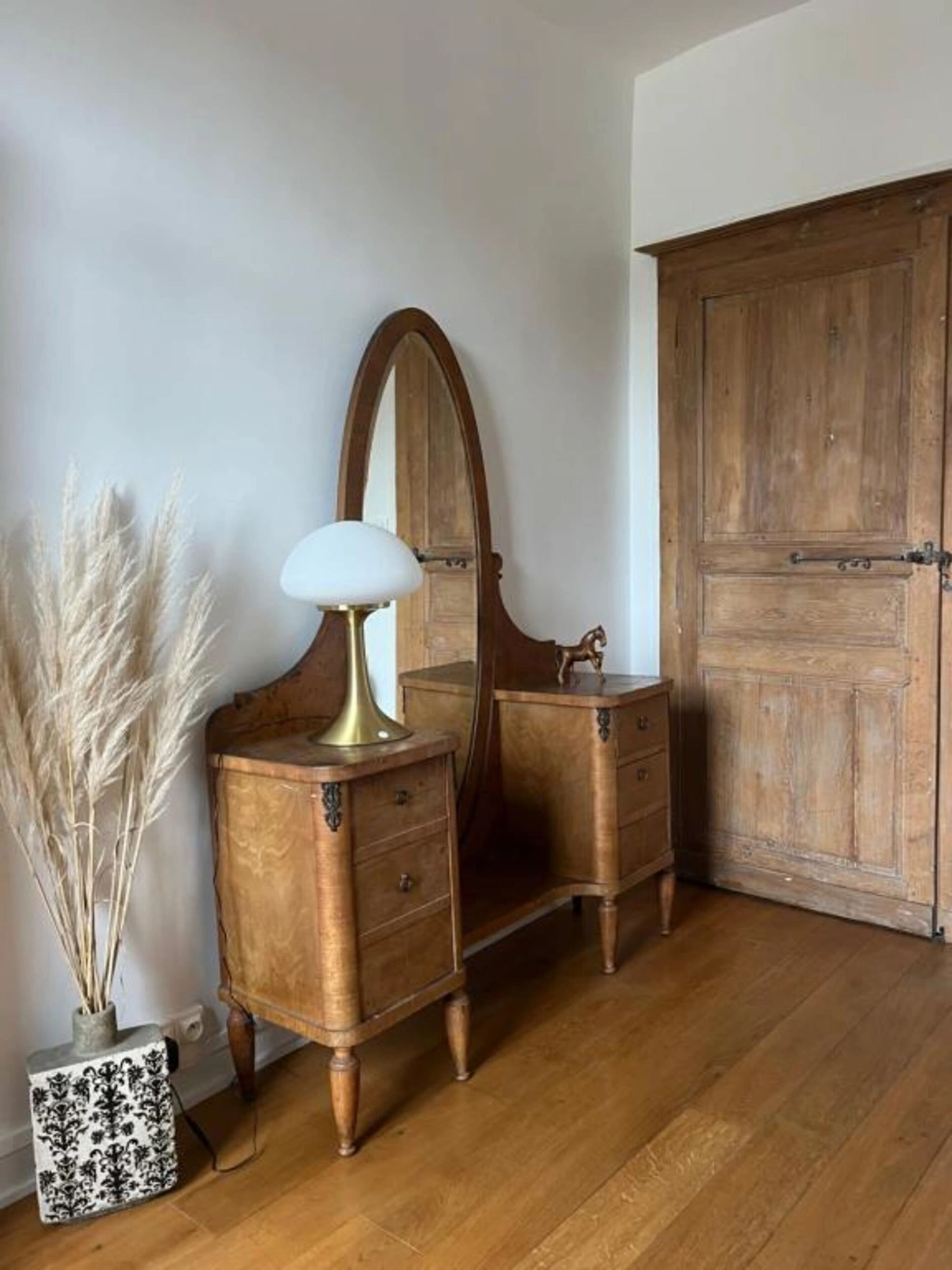 Une commode en bois avec un miroir, une lampe et une petite statue. On y trouve une grande porte en bois et un vase contenant des plantes séchées.