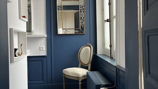 A narrow hallway with dark blue walls, a white radiator, a chair, and a mirror with decorative frame.