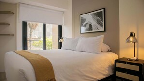 A neatly made bed with a white comforter and a brown throw blanket, a bedside table with a lamp, a framed picture on the wall, and a window with a view of greenery outside. - w* | Distinctive 1BR w/ Balcony in Barranco (Barranco)