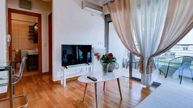 A modern living room with a flat-screen TV, a small white table with a vase of flowers, and a balcony with a chair. - Brand new flat in the amazing area of       Alimos (Alimos)