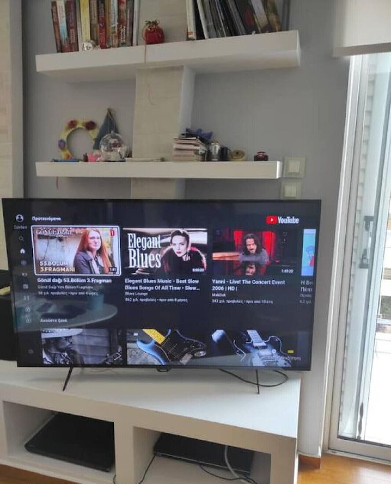 Ein Flachbildfernseher an einer weißen Unterhaltungsecke, darüber Regale mit Büchern und Dekorationsobjekten.