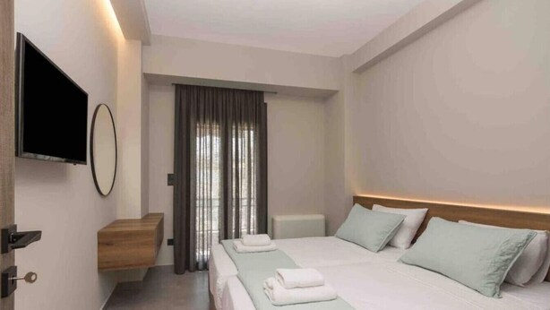 A modern hotel room with a bed, a TV mounted on the wall, a wooden side table, and a window with curtains. - Blue Pearl at Glufada sea side (Glifada)