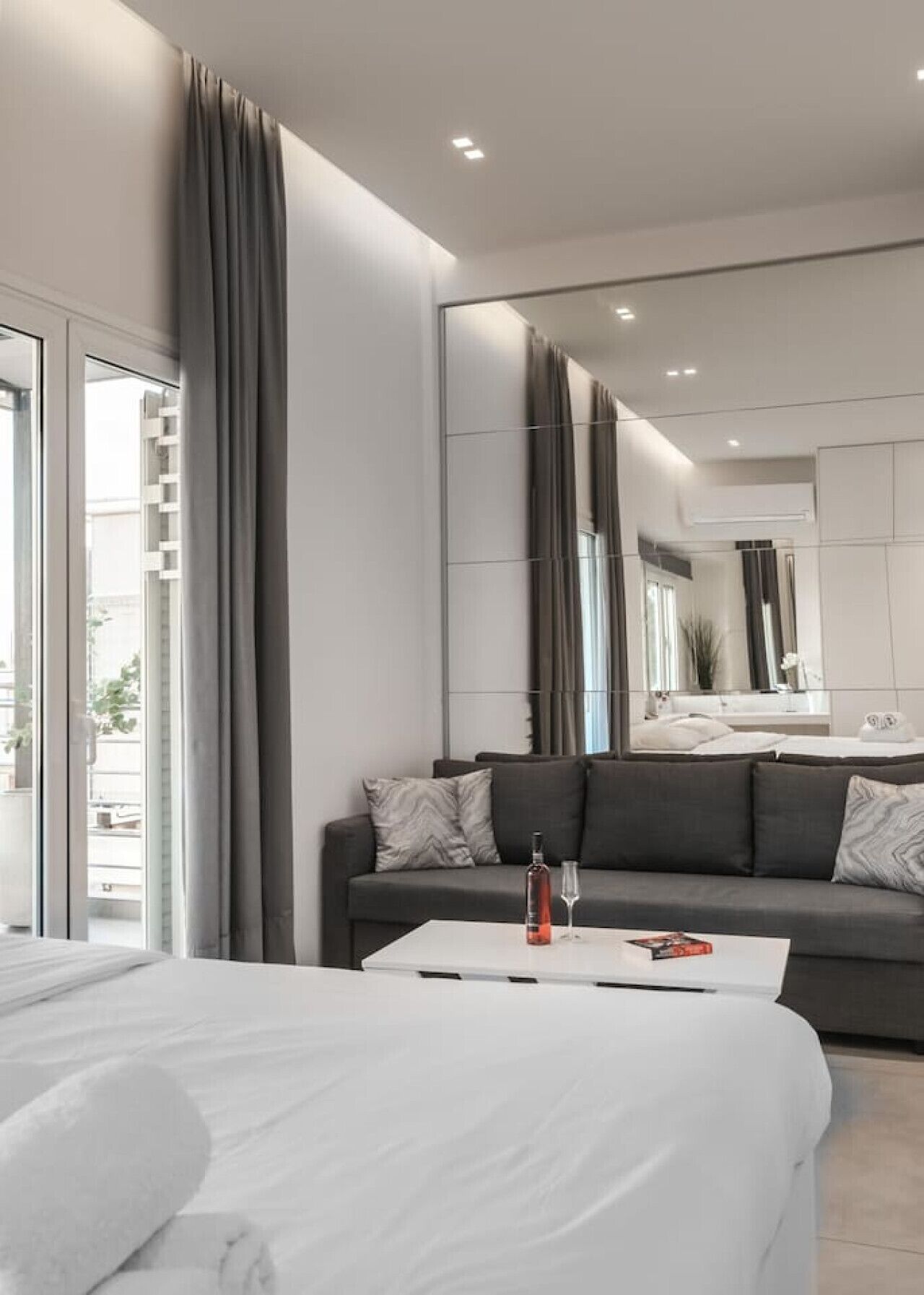Een moderne hotelkamer met een groot bed, een grijze bank, een witte salontafel en een grote spiegel.